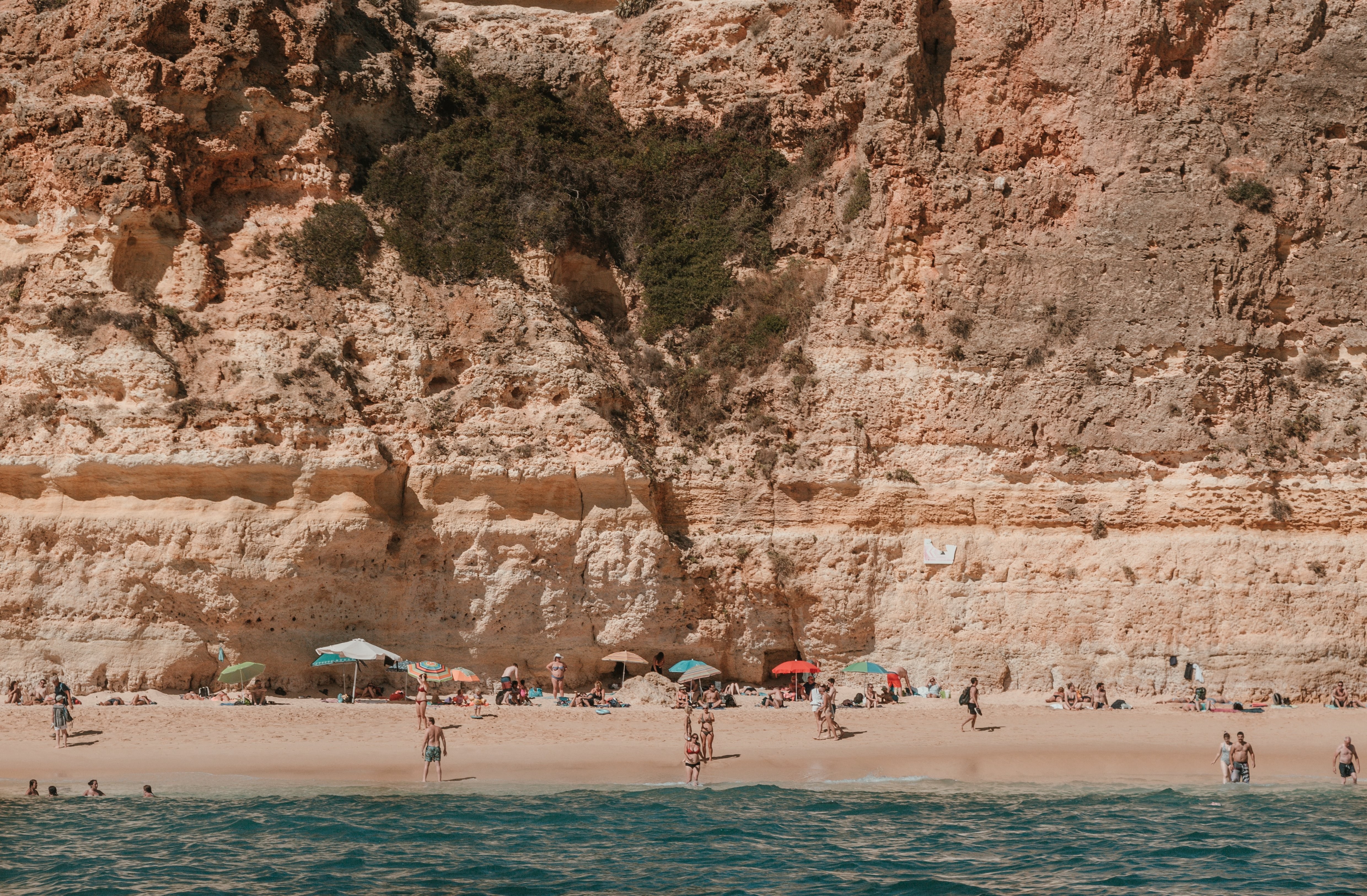 Sunny Beach Serenity: Bathers Relax Under Towering Rocky Cliffs – Stunning Photo