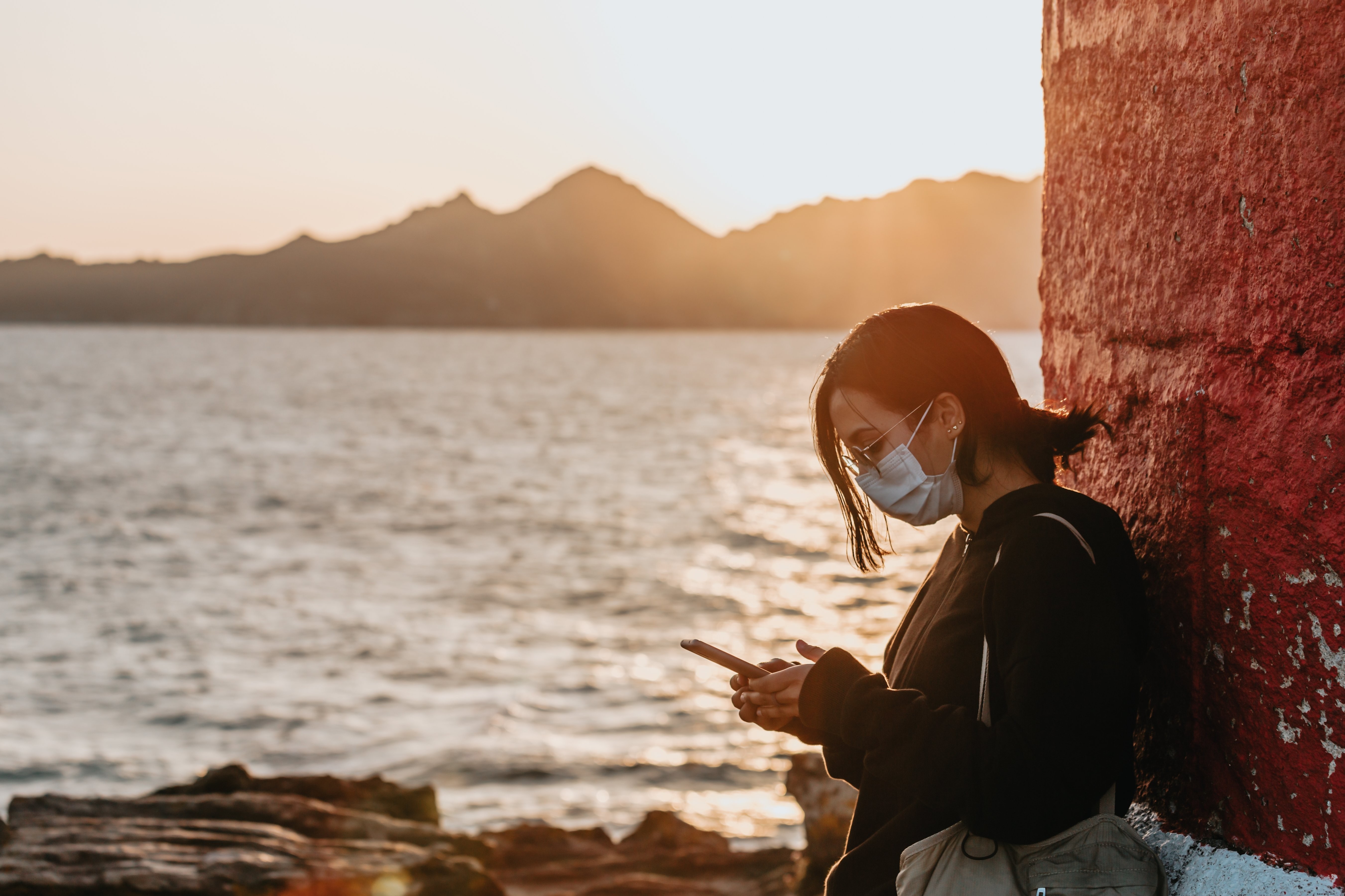 Woman in Face Mask Stands by Water, Checking Phone